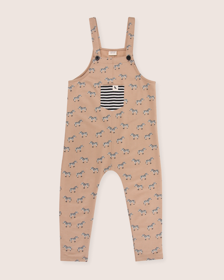 The Zebra Dungarees are eco-friendly kids overalls made from organic cotton, featuring a light brown base with a playful black and white zebra pattern. They include a front pocket with black and white stripes and button straps for casual style.