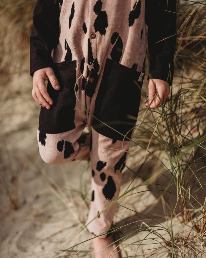 A child in Pony Velour Dungarees, made of organic fabric, stands on sandy ground with grass, wearing a cow-patterned jumpsuit with black sleeves and large black pockets.