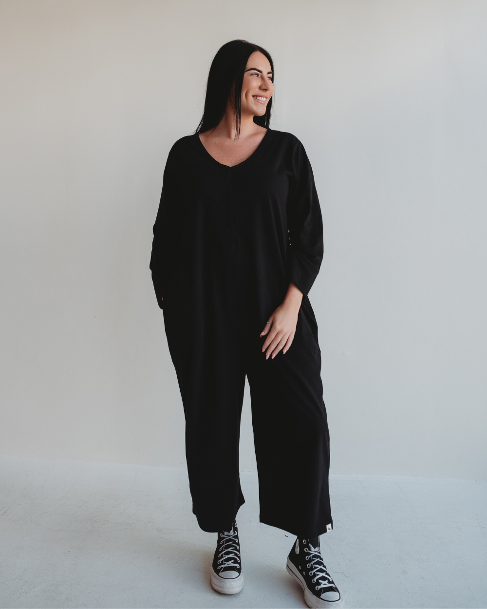 A person with long black hair stands against a plain white background, smiling and wearing Mai Black Dungarees made from organic cotton and black Converse sneakers.