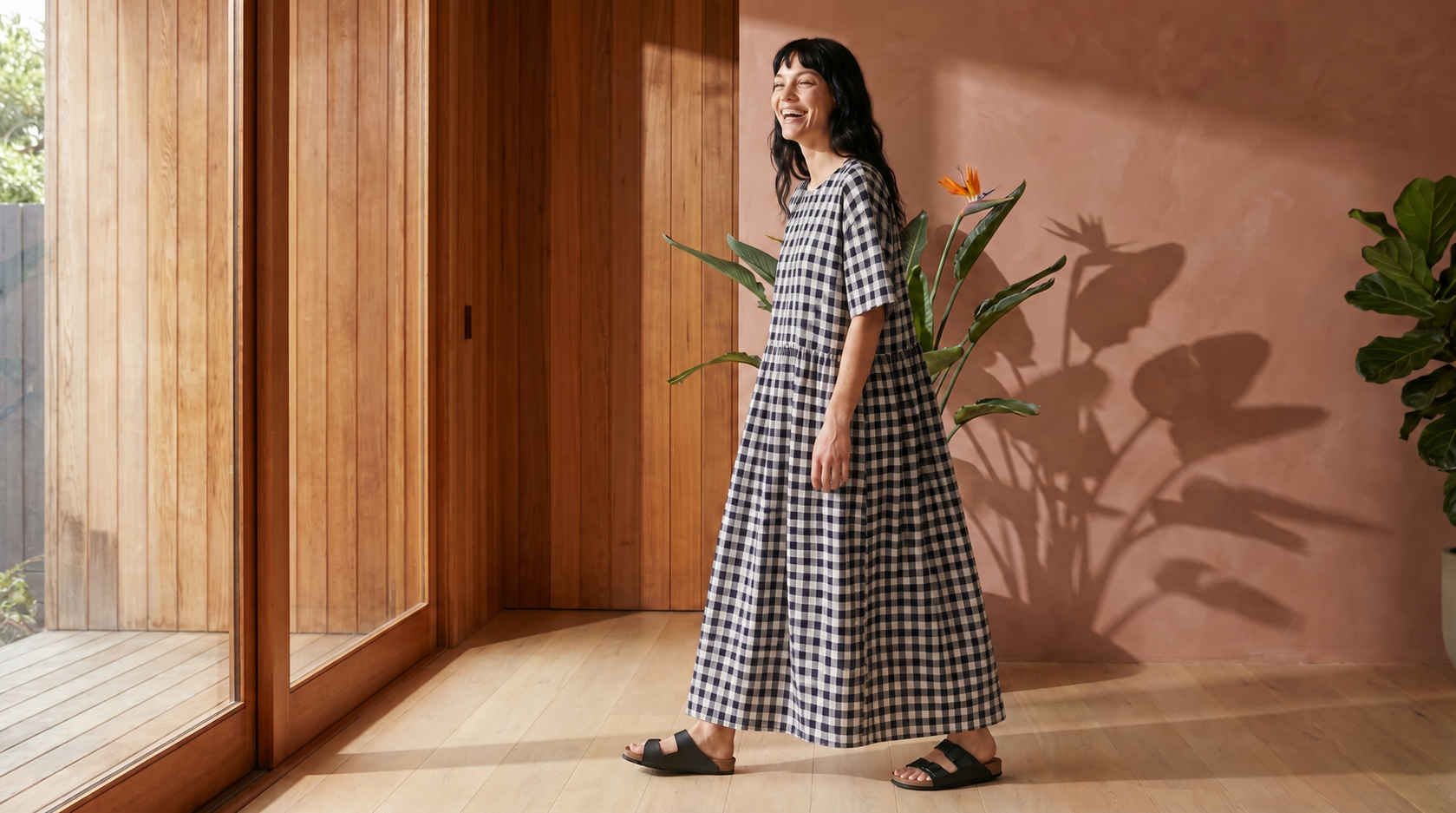 Woman in a checkered dress standing in a room with wooden walls and plants.