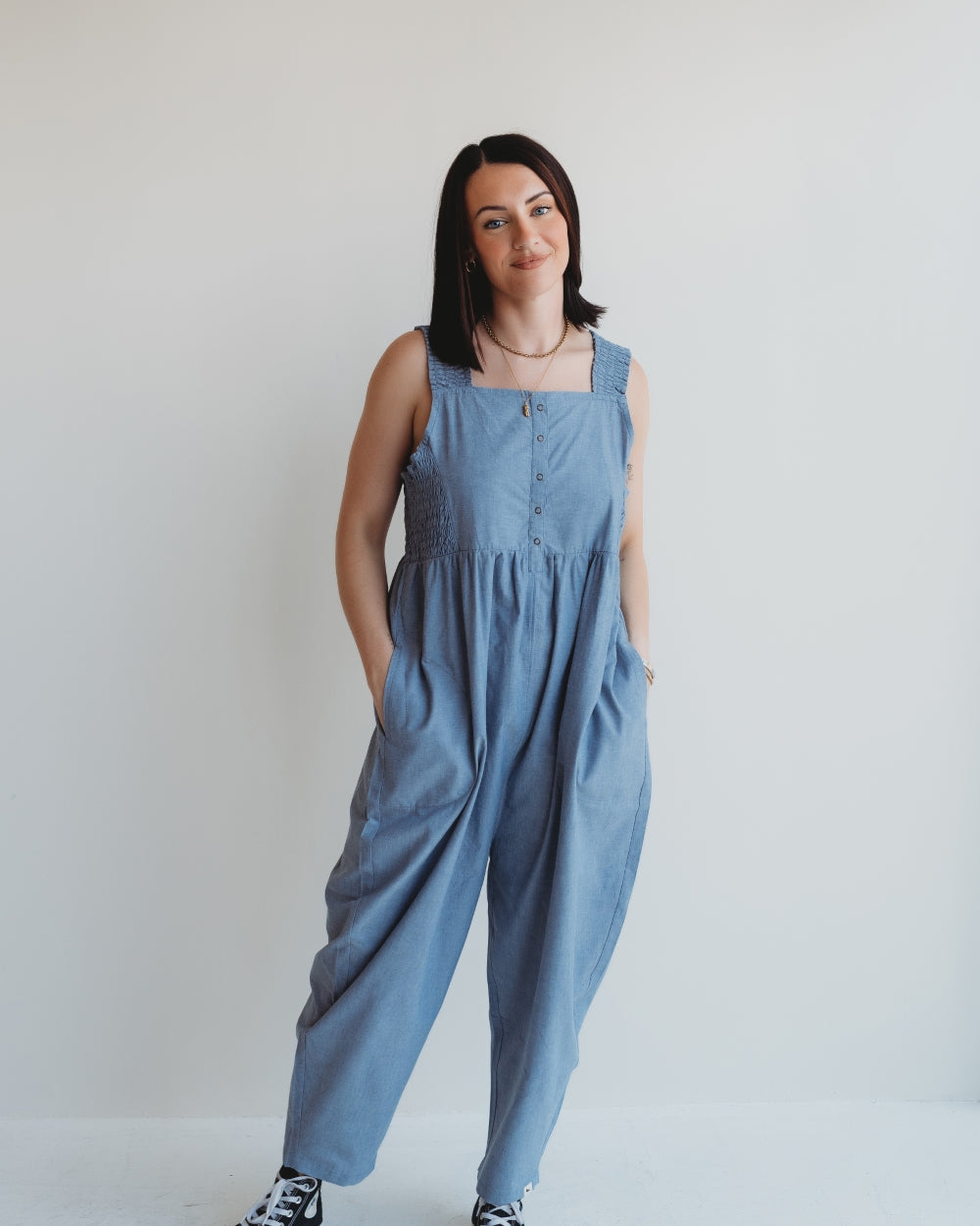 A person in Alice Chambray Dungarees stands against a plain white background with hands in pockets. Their shoulder-length dark hair pairs with black and white sneakers, highlighting effortless style with practical breastfeeding access.