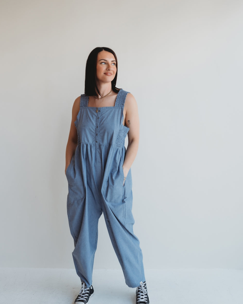 A woman in loose Alice Chambray Dungarees, crafted from organic chambray, stands against a plain white wall. Her long dark hair frames her smiling face as she gazes to the side. With hands tucked in her pockets, she completes her look with black and white sneakers.