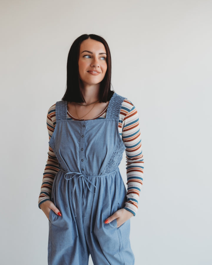 A person with long dark hair stands against a plain background, wearing a striped long-sleeve shirt under Alice Chambray Dungarees. Hands in pockets, they gaze to the side with a slight smile.