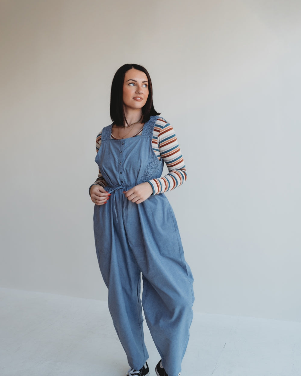 A woman with long dark hair stands against a white background, wearing Alice Chambray Dungarees over a striped long-sleeve shirt. She gazes to the side with a relaxed expression, showcasing effortless style and practical breastfeeding access.