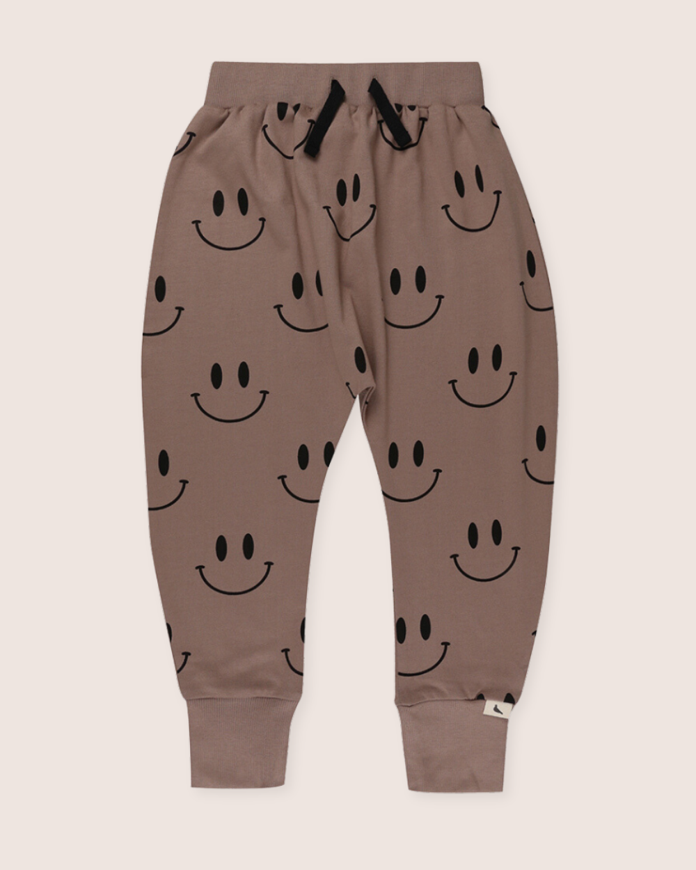 The Smiley Harems are brown jogger pants for kids made from organic cotton, featuring an elastic waistband, black drawstrings, cuffed ankles, and a playful Smiley Face print, all showcased against a light pink background.