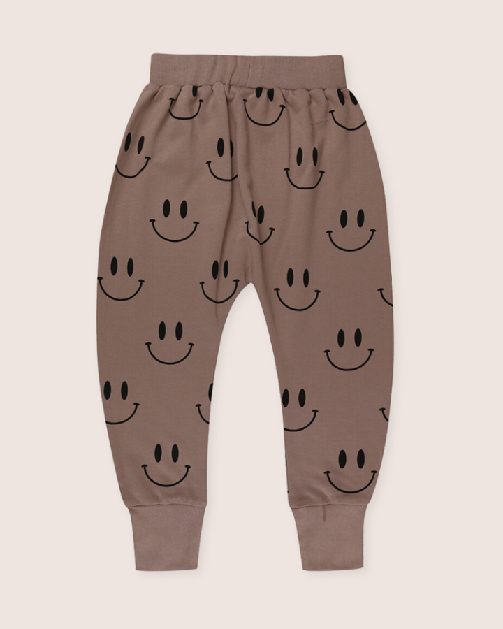 The Smiley Harems are brown kids joggers made from organic cotton, featuring an elastic waistband and cuffs with a playful smiley face print on a light, neutral background.