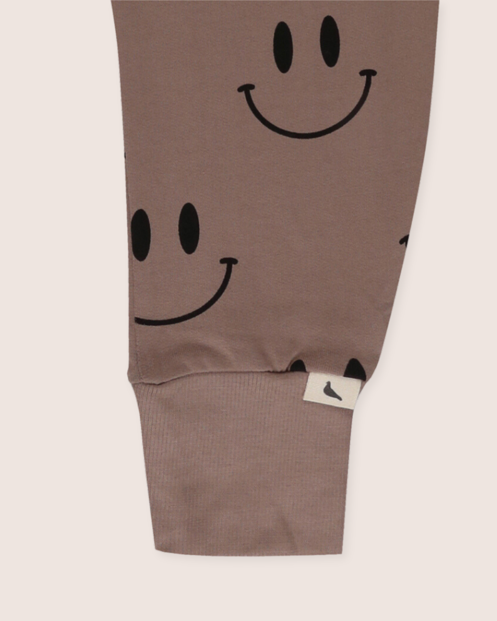 A close-up of the Smiley Harems jogger sleeve showcases a black Smiley Face print on organic cotton, with ribbed cuffs. A small white tag with a dark design is attached near the cuff.