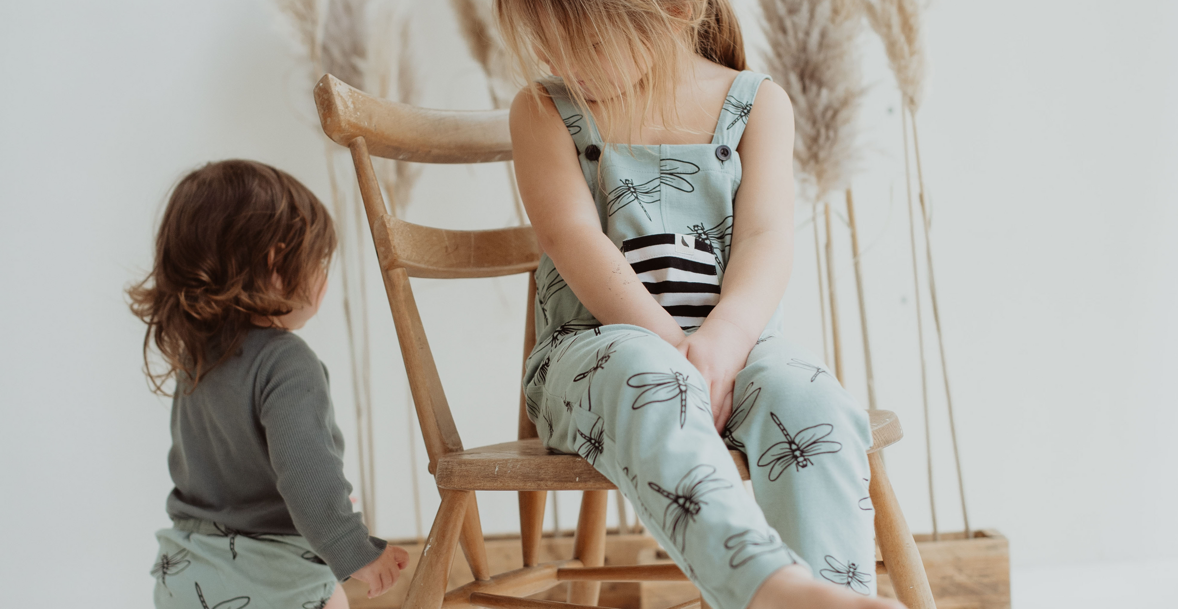 Child sits on a chair wearing turtledove dragonfly print organic cotton dungarees with stripe pocket. Other child wears the bloomer set of the same print. 