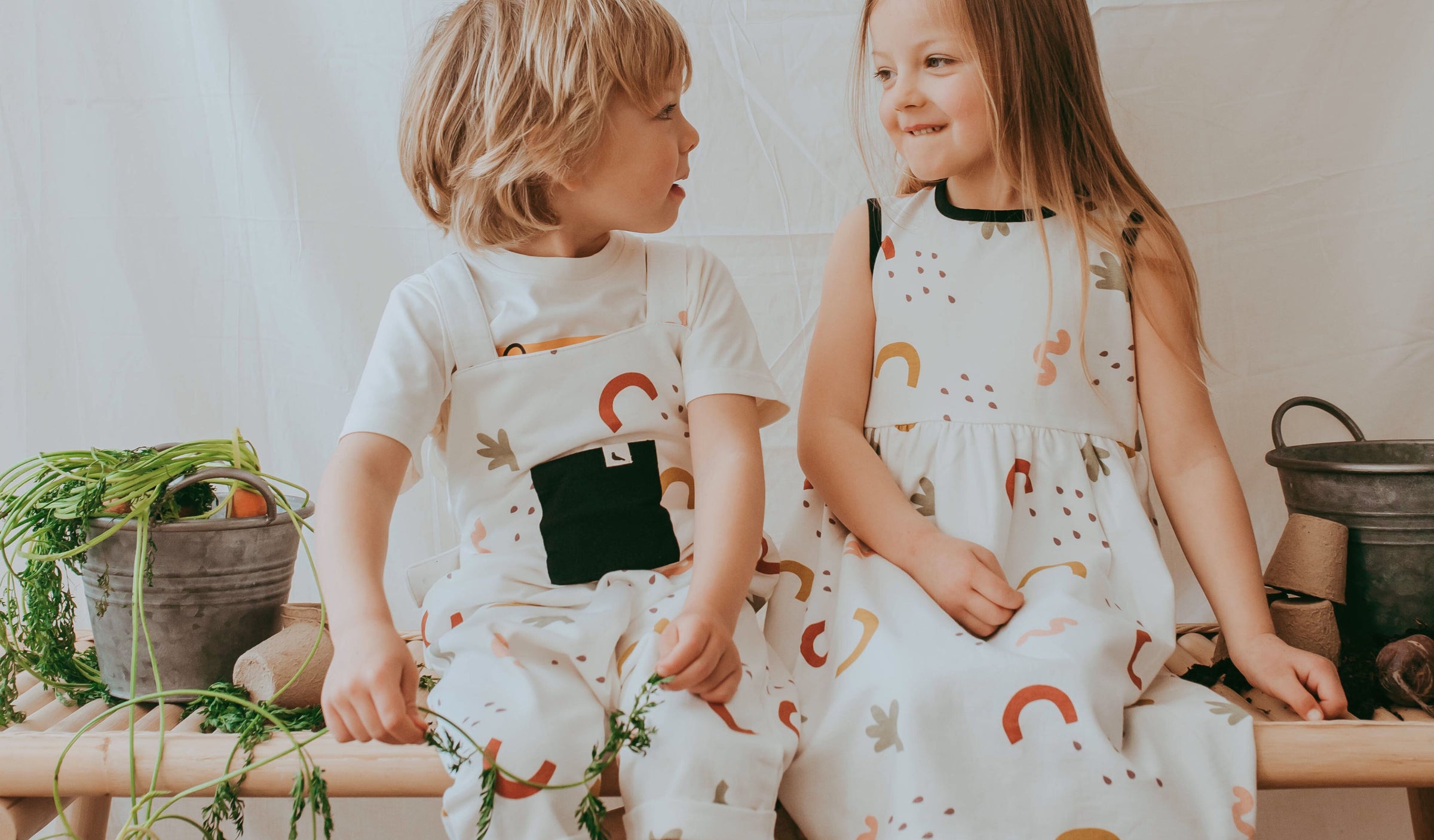 2 children sit together wearing TurtleDove organic cotton white silhouette print dress and dungarees. 