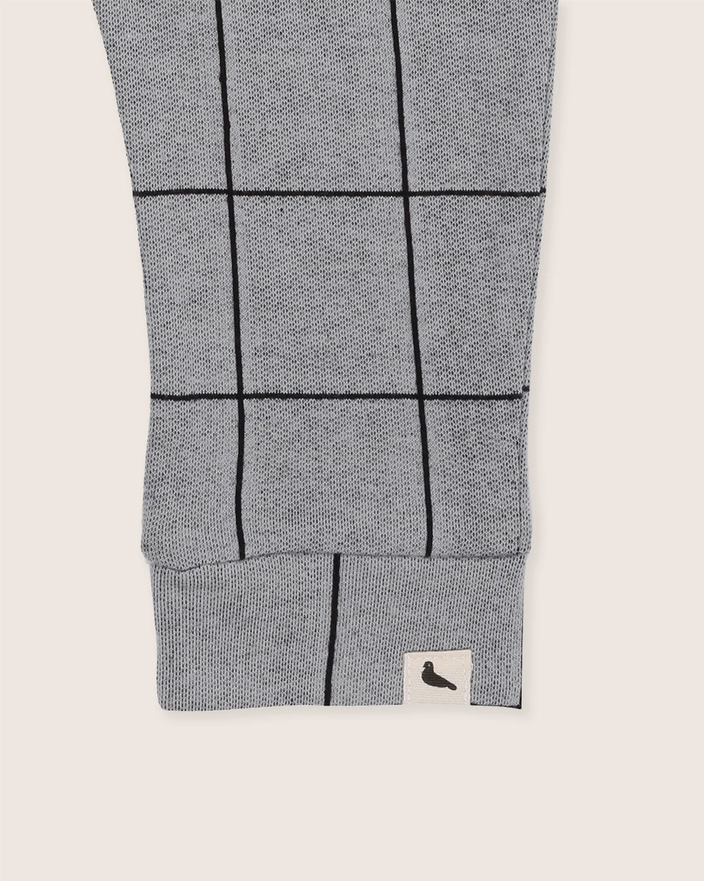 Close-up of a gray sweater sleeve showcasing a black Window Pane Check Print. The ribbed cuff and beige tag with a black bird detail enhance the design. Crafted from organic cotton, it pairs well with kids Window Pane Check Harem Joggers against an off-white background.