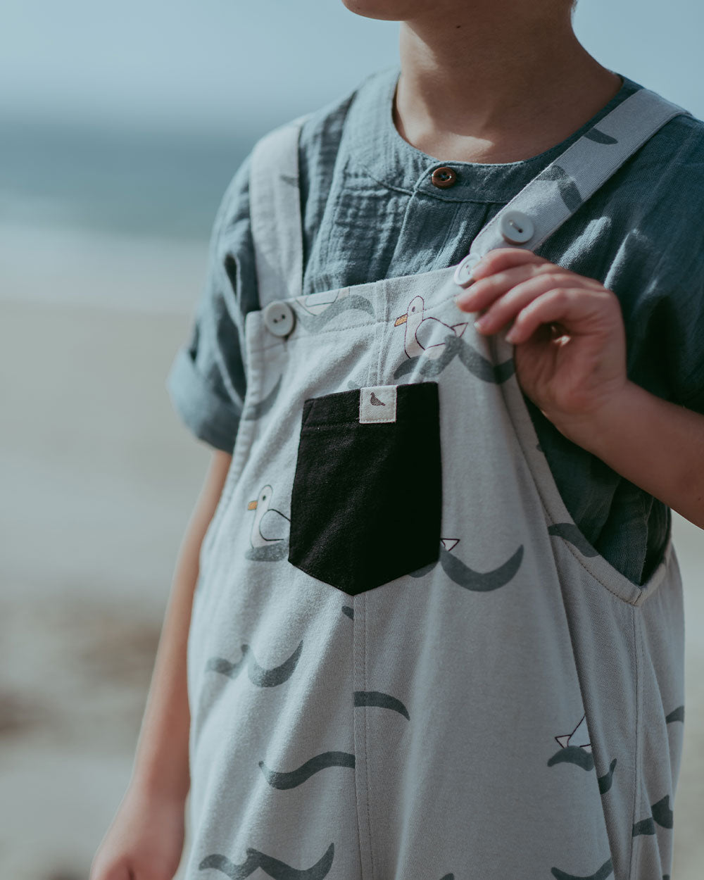 A child in beachwear dons Seagull Dungarees, blue, gender-neutral kids attire with seagull designs and a black pocket. Made from 100% GOTS organic cotton, this eco-friendly choice stands out against the blurred beach background.