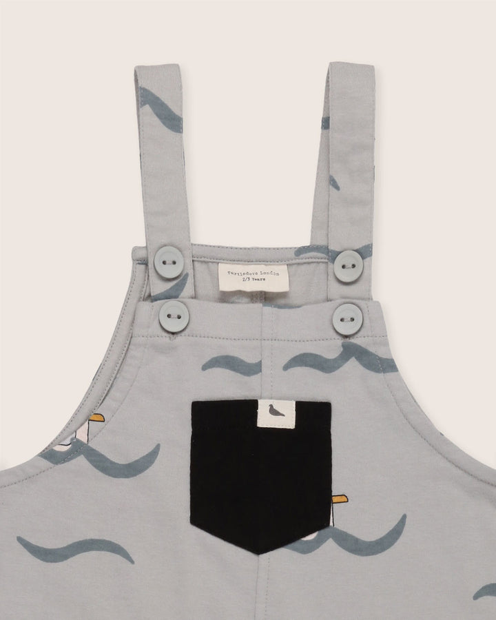 The Seagull Dungarees are gray, gender-neutral kids dungarees with seagull illustrations and wave designs. Made from 100% GOTS organic cotton, they have two buttons per strap and a black front pocket with a seagull design for an eco-friendly touch.