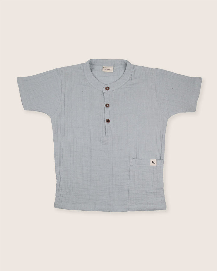 The Sky Gauze Shirt is a light blue short-sleeve top made from organic gauze fabric. It has a round collar, four front buttons, and features a small pocket with a subtle logo tag on the lower right side. Its textured, woven appearance makes it ideal for any sustainable capsule wardrobe.