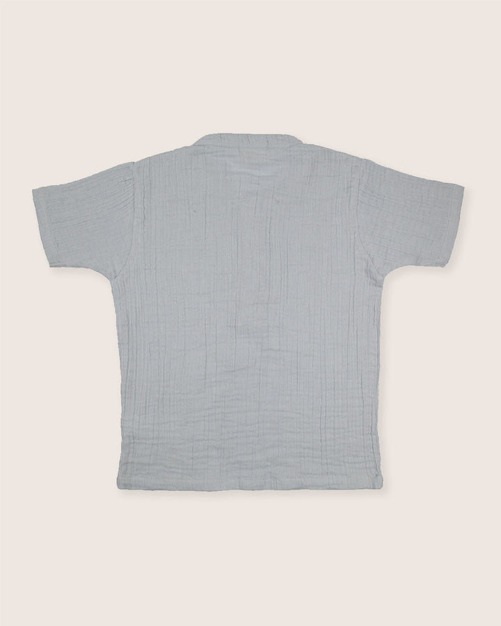 The Sky Gauze Shirt is a light grey, textured short-sleeve shirt with a mandarin collar. Made from organic gauze fabric, its shown from the back on an off-white background—ideal for a sustainable capsule wardrobe.