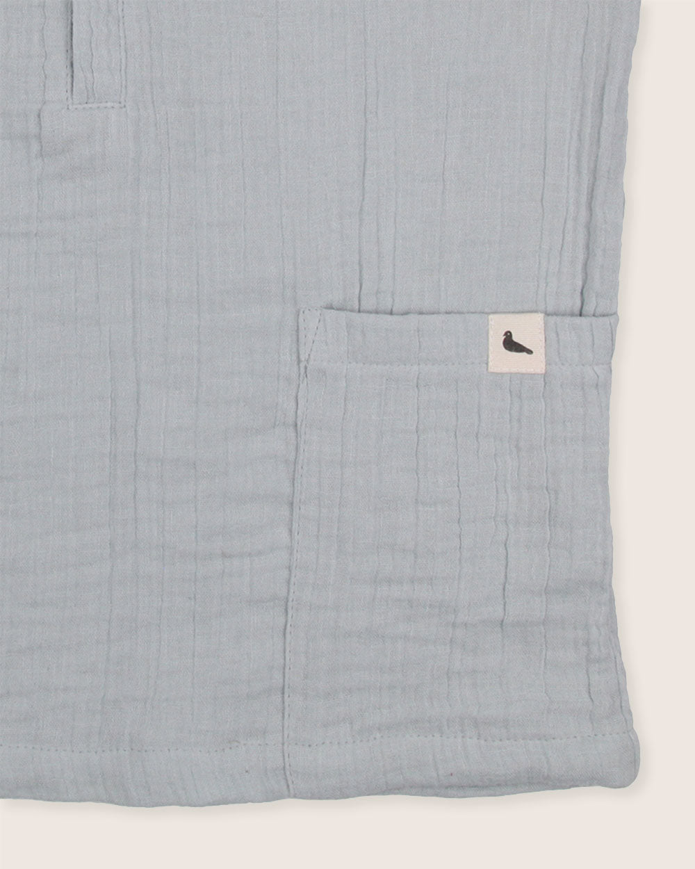 A close-up of the Sky Gauze Shirt showcases its textured light gray organic fabric with a small pocket. A white tag with a black bird silhouette is sewn onto the pocket, making it ideal for your sustainable capsule wardrobe.