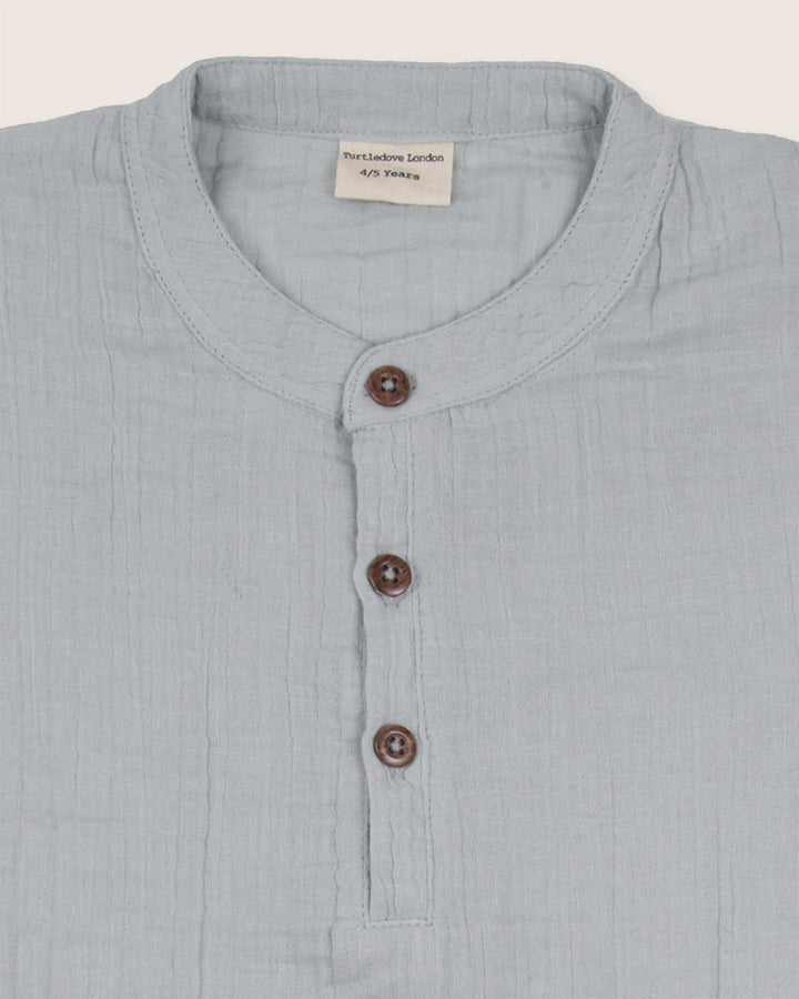 The Sky Gauze Shirt is a light blue childrens shirt made from organic gauze fabric, featuring a textured appearance and a mandarin collar. It includes three brown buttons and an inside collar label, ideal for a sustainable capsule wardrobe.