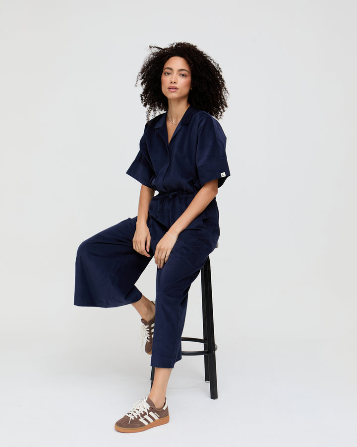 Woman wearing a navy jumpsuit sitting on a black stool against a white background