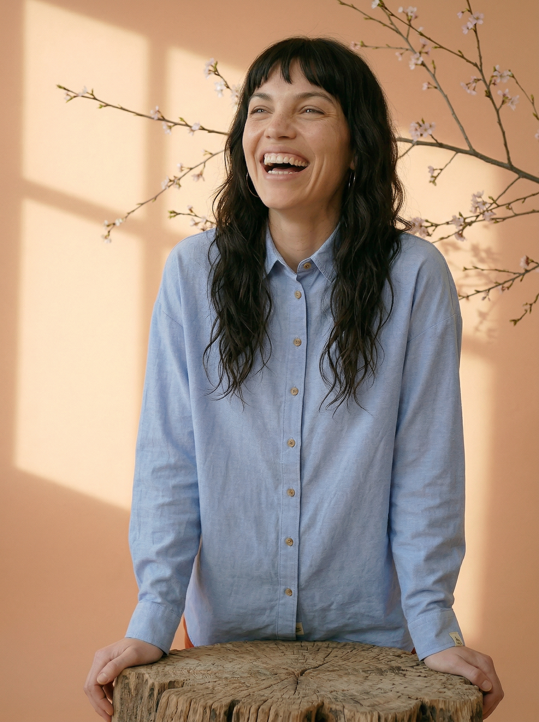 Woman in a light blue shirt standing behind a wooden stool with a soft peach background and floral branches.