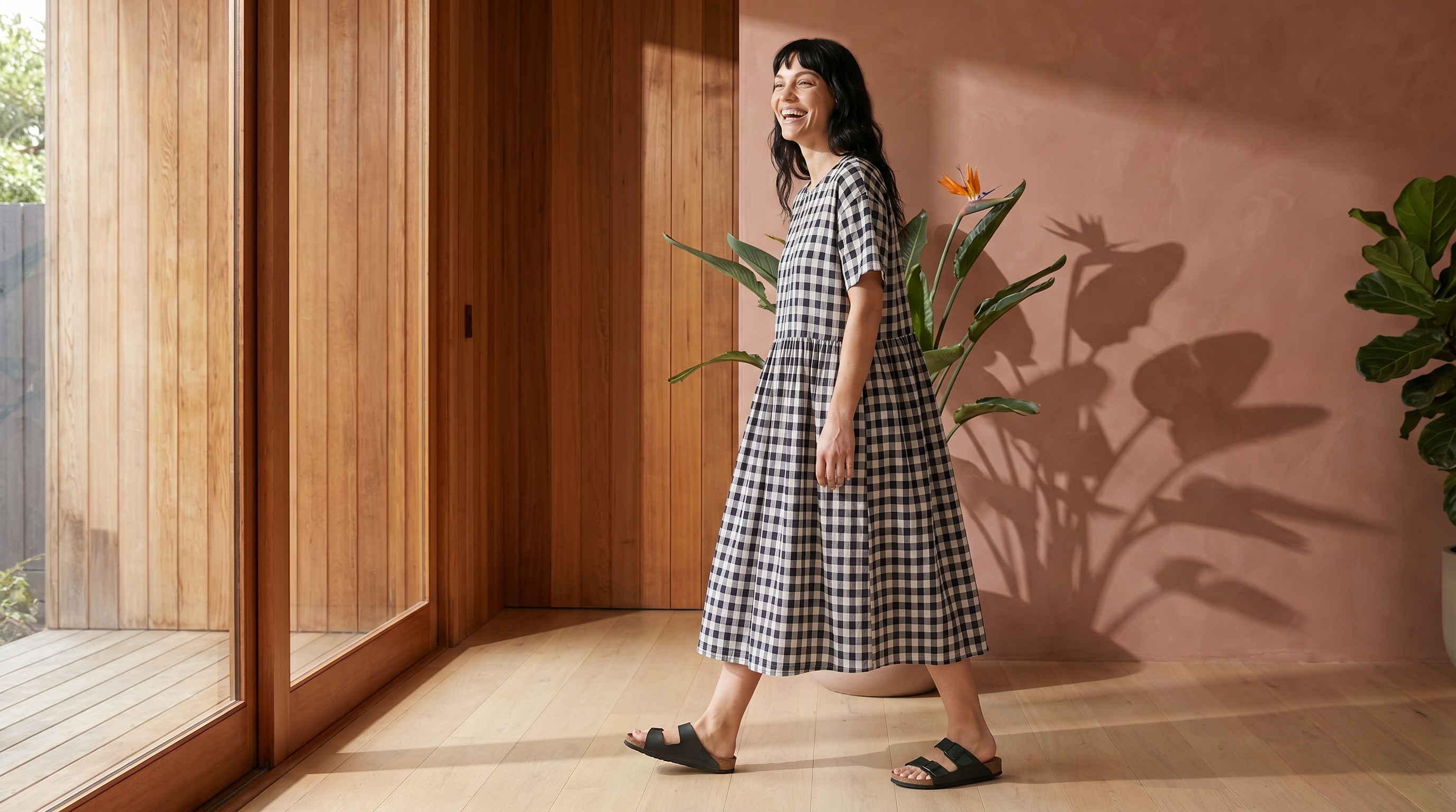 Woman in a checkered dress standing in a room with wooden walls and plants.
