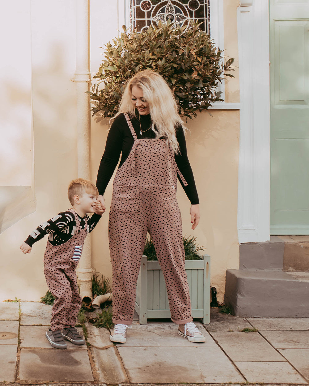 A woman and child smile on a sidewalk before a house with a pale yellow wall and green door. Wearing matching Lila Confetti Dungarees crafted from organic cotton with black designs, the woman warmly holds the childs hand.