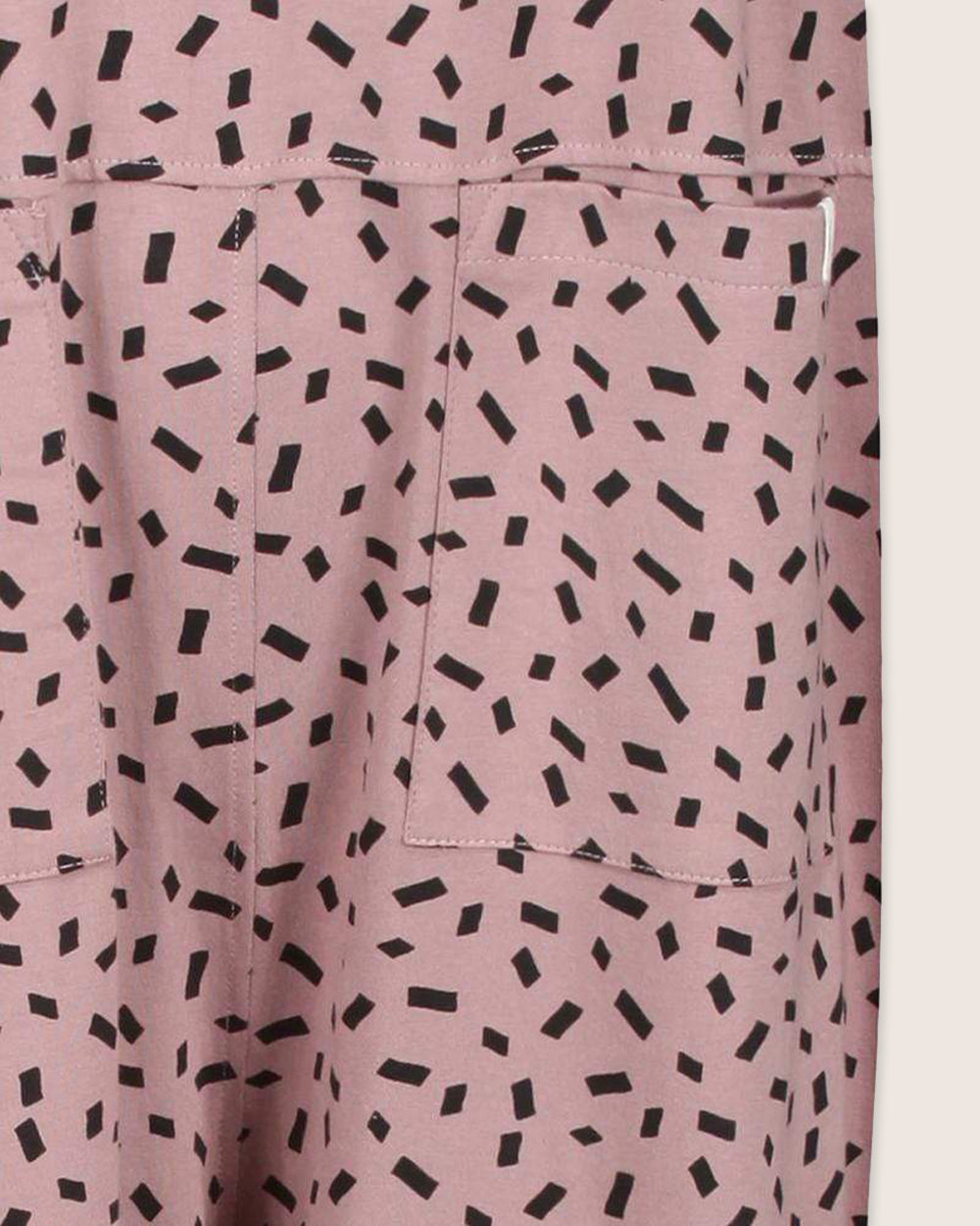 Close-up of pink Lila Confetti Dungarees with black rectangular patterns. Made from organic cotton, these dungarees feature two pockets with visible stitching on an off-white background, representing sustainable fashion.