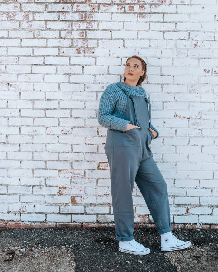 A person stands against a white brick wall, wearing a light blue sweater with Lila Dark Blue Dungarees and white sneakers. They have their hands in their pockets and gaze upwards.